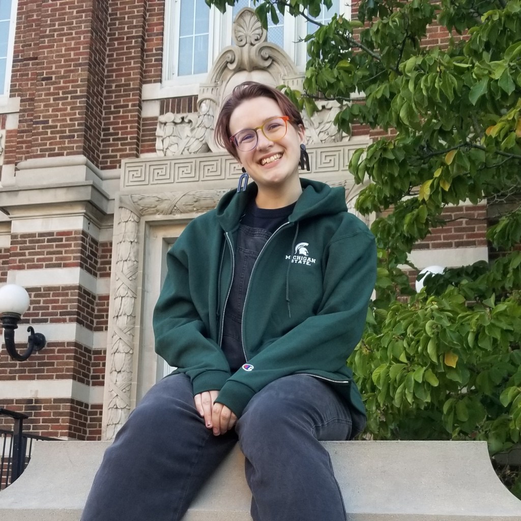 Image of Faye, a white person with short brown hair and rainbow glasses, sitting in front of a brick building, wearing a green Michigan State sweatshirt